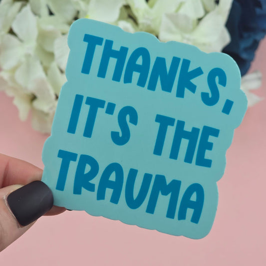 Blue sticker with text 'Thanks, it's the trauma' held by a hand against a pink background