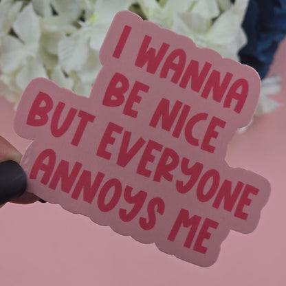 Sticker with red text 'I WANNA BE NICE BUT EVERYONE ANNOYS ME' held in front of white flowers.