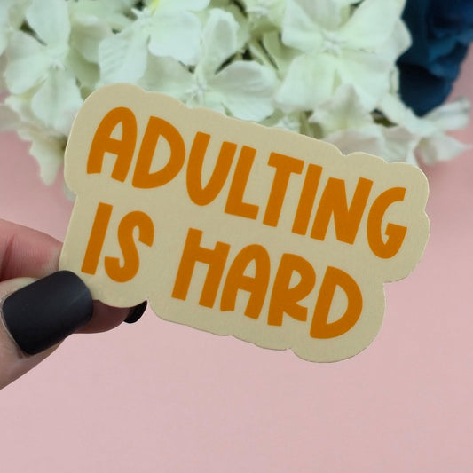 Sticker with 'ADULTING IS HARD' text held over white flowers against a pink background